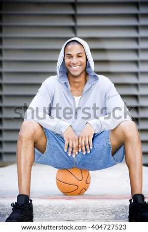 Portrait of a smiling young African American man in a grey hooded top sitting on a basketball.