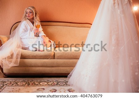 Beautiful blonde bride in robe posing with champagne near dress