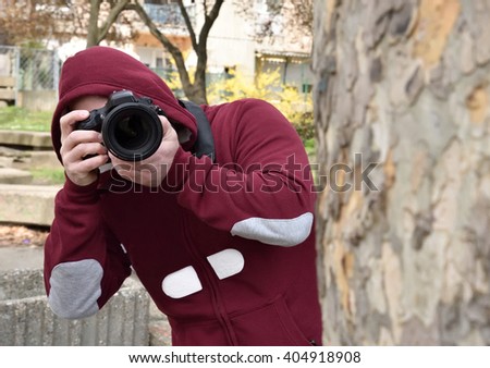 Mysterious paparazzi photographer with the hood on his head lurking behind the tree