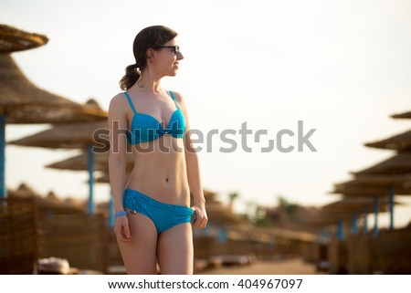 Young slim happy smiling woman in sunglasses standing in relaxed pose on empty beach in front of umbrellas, relaxing in sunlight on tropical sea resort