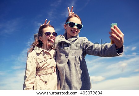 people, children, technology, friends and friendship concept - happy girls with smartphone taking selfie and making funny faces outdoors