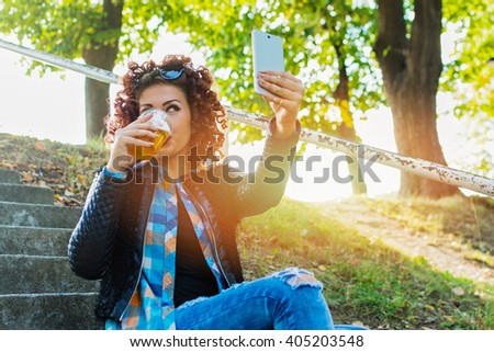 Modern young woman in casual clothes, with red curly hair, taking a selfie in park in spring and drinking beer. Horizontal, vibrant colors, natural light, mild retouch.
