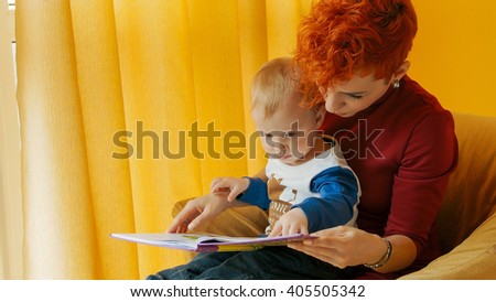 Mother with baby sitting in chair and reading story with pictures. Little boy looks into book and shows finger in book. Family, early development, activity, learning. Cute kid, child play with mom.