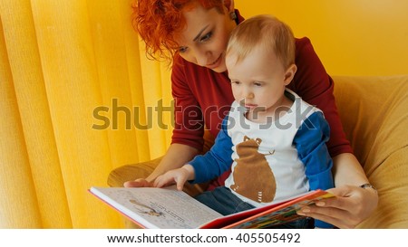 Mother with baby sitting in chair and reading story with pictures. Little boy looks into book and shows finger in book. Family, early development, activity, learning. Cute kid, child play with mom.