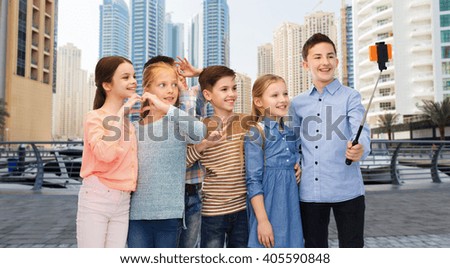 childhood, friendship, technology and people concept - group of happy children talking picture by smartphone on selfie stick over dubai city street background