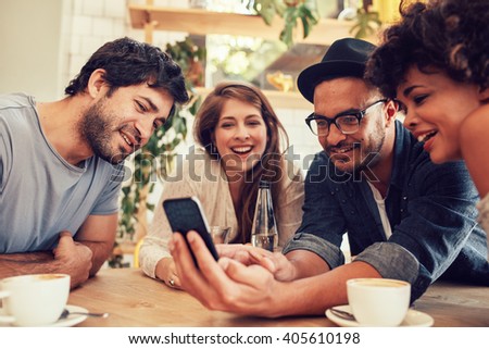 Group of young people sitting in a cafe and looking at the photos on smart phone. Young men and women meeting at cafe table and using cell phone