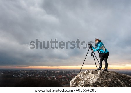 Young female photographer is standing with her camera on tripod on the big rock at city overview point. Woman is taking picture of the cityscape in the evening