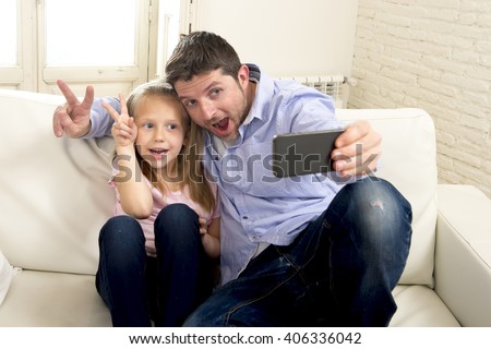 young happy man having fun with his little cute blond daughter taking selfie photo with mobile phone enjoying together at home sofa couch in father and little girl self portrait picture concept