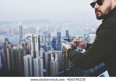 Closely image of a bearded hipster guy in fashionable sunglasses with mobile phone in hand is looking away, while is standing on building roof against blurred business district skyscrapers in New York