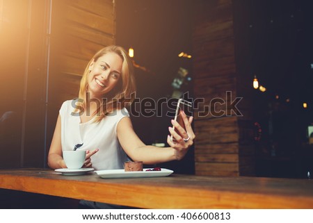 Charming happy hipster girl photographing herself on cell telephone camera while relaxing in modern coffee shop, pretty woman with cute smile making self photo on her smart phone during rest in cafe 