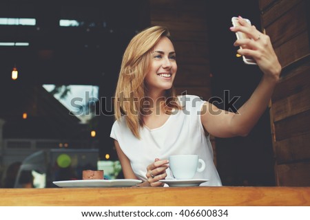 Happy smiling hipster girl making self portrait with mobile phone camera while sitting in modern coffee shop, cheerful woman posing while photographing herself for social network picture, copy space