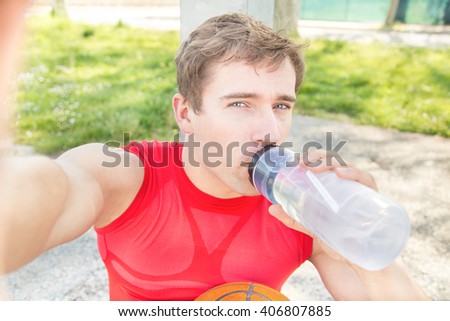 Sitting guy in a basketball court and take a selfie after match with friends. Sport man drinking from water bottle. Young man finish do sport and make himself picture. Fitness jogging workout concept.