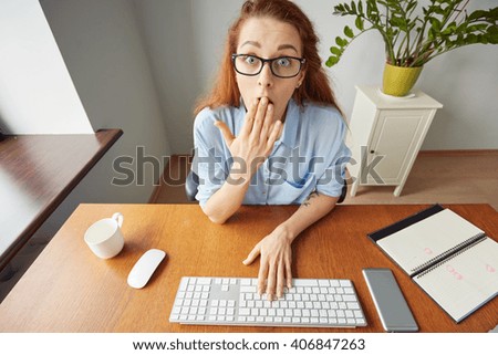 Top view portrait of dissatisfied young female office worker looking unhappily at the camera, leaning on her elbow while sitting at wooden table in front of computer screen during hard working day  