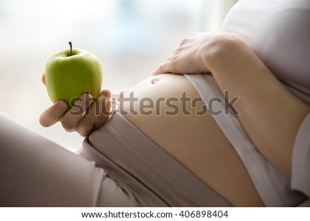 Close-up of torso of young pregnant model holding green apple. Future mom expecting baby eating fruits at home. Closeup of belly. Healthy eating concept