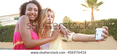 Panoramic portrait of ethnically diverse teenager girls posing together pulling faces using a smart phone to take selfies on sunset suburban street, outdoors lifestyle. Young people using technology.