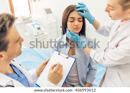 Beautiful young woman is sitting with closed eyes in doctors office, two doctors are examining her face and making notes Royalty-Free Stock Photo #406993612