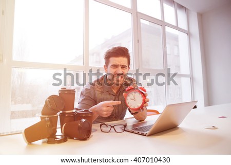 Tired student holding red alarm-clock in library demonstrating that it is time for taking exams. Astonished man looking frighnetened. Toned picture. 