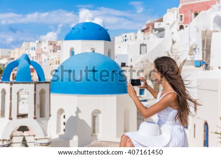 Europe travel woman taking photo photographing using smart phone in Oia, Santorini, Greece. Famous blue domes on white church in village. Young asian tourist taking pictures on smartphone on holidays