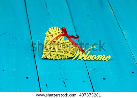Bright yellow wicker heart with red ribbon with the word Welcome on antique rustic teal blue wood background; Valentines Day, love and home concept background with painted copy space