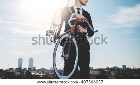 Professional businessman wearing black suit and standing with modern orange bicycle, panoramic view of the city in the background, flare light, outdoors