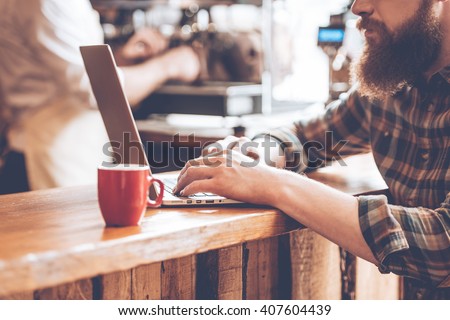 Writing post from cafe. Close-up part of young bearded man using his laptop while sitting at bar counter at cafe with barista at the background Royalty-Free Stock Photo #407604439