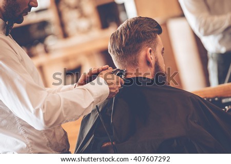 Perfect  trim. Rear view close-up of young bearded man getting haircut by hairdresser with electric razor while sitting in chair at barbershop Royalty-Free Stock Photo #407607292
