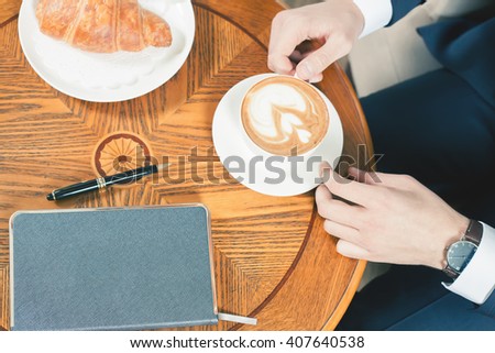 Close-up image of coffee break with a cap of coffee and croissant. Businessman in a jacket sits at the table at office. Good Morning. Cafe, restaurant