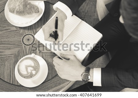 Close-up image of coffee break with a cap of coffee and croissant. Black and white, retro. Businessman writes something in notebook, office. Good Morning monday. Cafe, restaurant