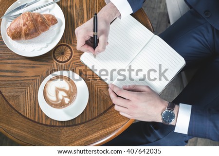 Close-up image of coffee break with a cap of coffee and croissant. Businessman writes something in notebook, office. Good Morning. Cafe, restaurant