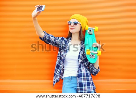 Pretty cool girl in sunglasses with skateboard taking picture self portrait on smartphone over orange background