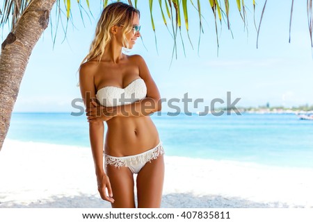 Young woman wearing white bikini posing under palm tree over sea view at tropical beach