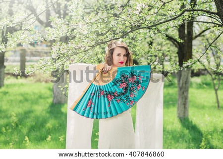 young girl in flowering garden in bloom and flying white dress, emotion on face unusual sight smile sunlight outdoor with large fan in Japanese style. Translation of the text  fantastic spring