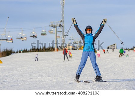 Full length portrait of happy young female skier on a sunny day at ski resort against ski-lift. Girl is standing on a ski slope with raised arms as sign of success. Winter vacation.