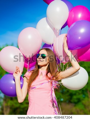 Beautiful fun woman holding multicolored helium balloons. Has smiling face, long hair, clothed pink dress, sunglasses. Has slim body. Portrait in green forest. Sunny day and blue sky. Close up.