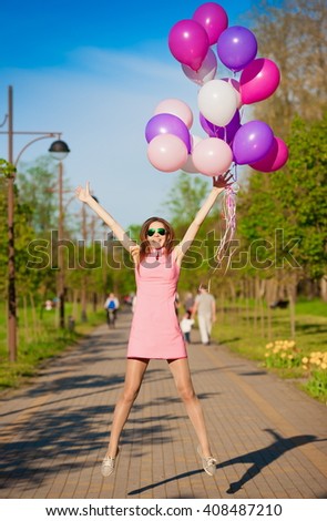Beautiful fun woman jumping with helium balloons. Has smiling face, long hair and legs, clothed pink dress, sunglasses. Has slim body. Portrait in city park. Sunny day and blue sky.