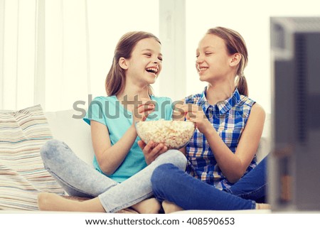 people, children, friends and friendship concept - two happy little girls watching tv, laughing and eating popcorn at home
