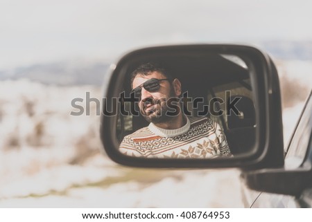 Young man driving the car is seen reflected in the side mirror with mountains in the background. Travel and adventure concept. Toned picture
