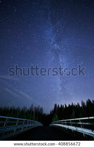 A night sky full of stars and visible milky way with a bridge on foreground. Road leading to dark forest.