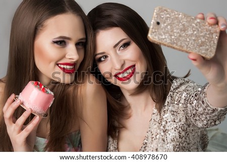 Beautiful girls eating cakes and making a photo of themselves. Red lips.  Smiling ladies. Fashion Brunette and blonde Portrait on a silver background. Gorgeous Woman Face. Ladies on the party. 
