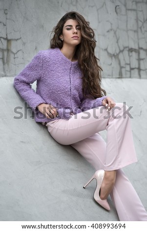 Attractive girl with beautiful hair sits on the concrete wall and looks into the camera. She wears violet jacket, pink pants and pink shoes. She holds right hand on her hip, left hand is on the knee