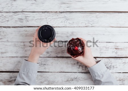 businessman hold the  chocolate muffin with cherry and coffee cup, hands detail on a wooden office table. the work place on background. business concept. desktop top view, unrecognizable people