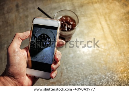 Hand taking picture of iced coffee americano on metal table surface with mobile phone with golden lighting
