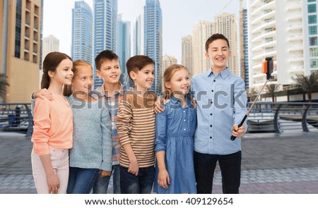 childhood, friendship, technology and people concept - group of happy children talking picture by smartphone on selfie stick over dubai city street background