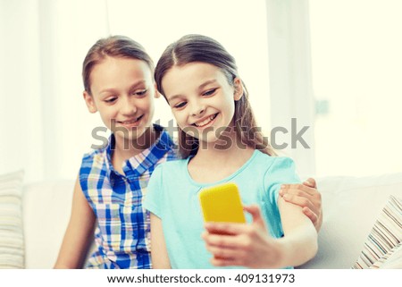 people, children, technology, friends and friendship concept - happy little girls sitting on sofa and taking selfie with smartphone and hugging at home