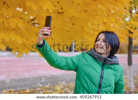 Smiling young woman in thick winter coat taking picture of herself at park with camera phone in front of yellow tree leaves in background