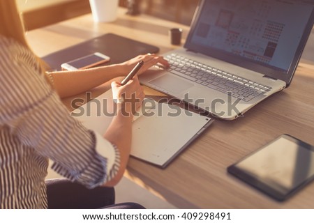 Female designer using laptop, sketching at blank notepad. Woman hand writing in notebook on wooden desk. Royalty-Free Stock Photo #409298491