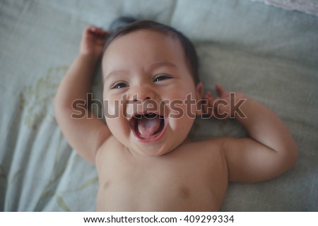 Close up of cute 10 month old mixed race baby boy lies happily on a bed. Dark natural indoor tones, window lighting only Royalty-Free Stock Photo #409299334