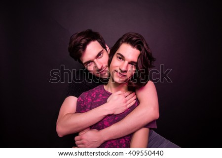 Cute beautiful young gay male couple. Two young homosexual guys hugging. Studio shot, black background, colorfully lit with colored lightning gels.