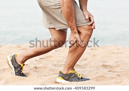 Muscle injury. Man with sprain thigh muscles. Athlete in sports shorts clutching his thigh muscles after pulling or straining them while jogging on the beach. Royalty-Free Stock Photo #409583299