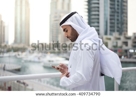 Middle Eastern Emirati Man in traditional clothing called kandura is standing on terrace in Dubai Marina and checking his mobile. Royalty-Free Stock Photo #409734790